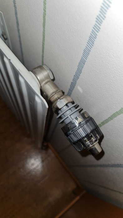 Termostatventil på ett radiatorselement med en fast insexskruv synlig undertill, mot en vägg med tapet.