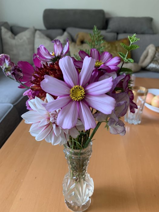 En bukett med blandade rosa och lila blommor i en glasvas på ett bord med en soffa i bakgrunden.