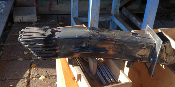 Svetsad prototyp av en pelletsbrännare med zigzagmönster och sluten korg, ligger på träplankor utomhus.