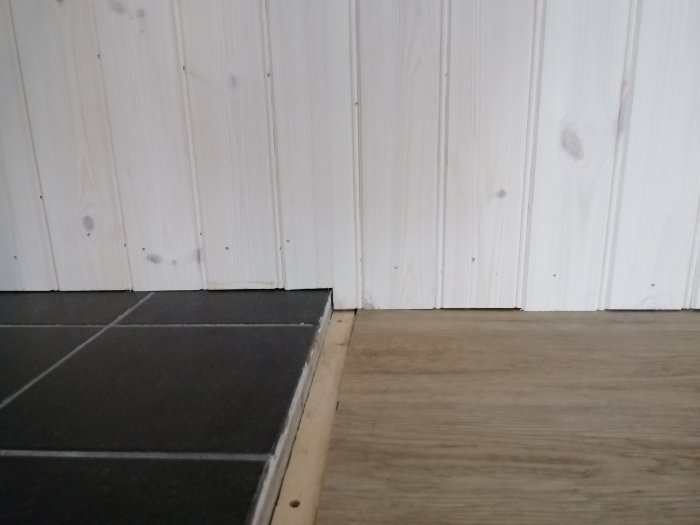 Vit träpanelvägg möter golv med klinkerplattor och träimitation.