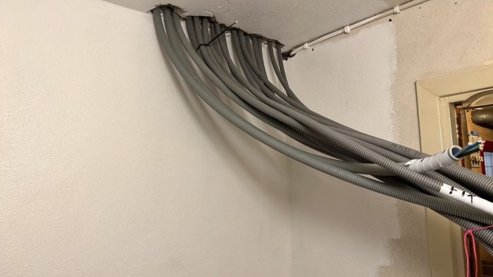 Grå elrör bundna tillsammans leder längs en vit vägg och hörn som en del av ett elektriskt installationsprojekt.