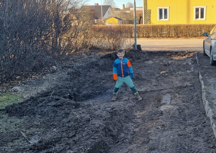 Ett barn står leende på grävd jord vid en blivande garageuppfart, med hål och spår från grävarbetet synliga.