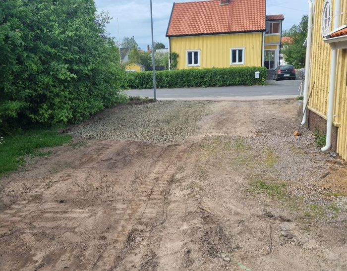 En nyanlagd garageuppfart med grus och delvis planmarkerad yta bredvid en gul husvägg och en nedklippt häck.