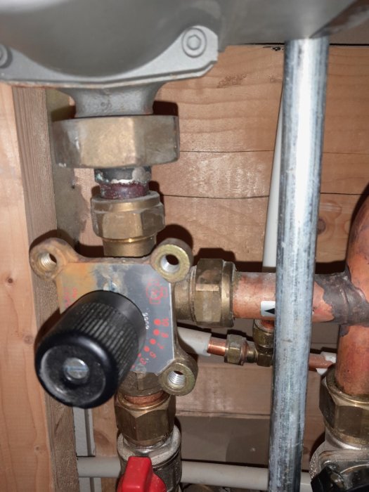 VVS-installation med kopparledningar och ventil med svart ratt mot en trävägg.