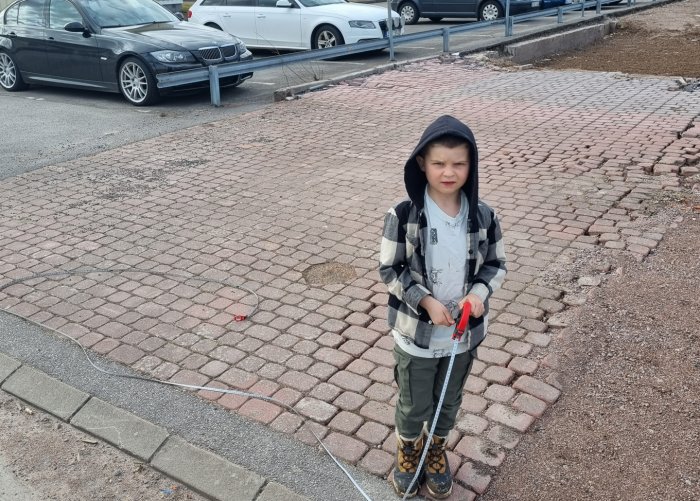 Barn står på marksten, håller i mätverktyg, med parkerade bilar och utgrävningsområde i bakgrunden.