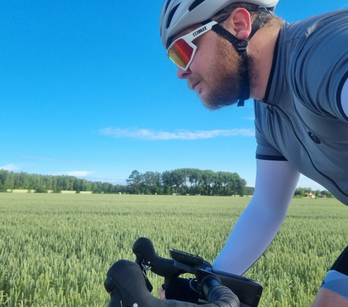 Cyklist i hjälm och solglasögon cyklar bredvid ett grönt fält under solig himmel.