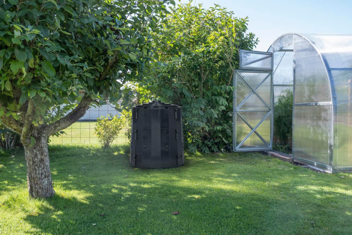 Trädgård med fruktträd, svart kompostbehållare och genomskinligt växthus på gräsmatta.
