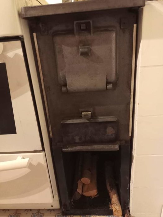 En gammal vedeldad kamin i svart stål med vedfack och en stängd lucka, placerad vid en vit toalett.