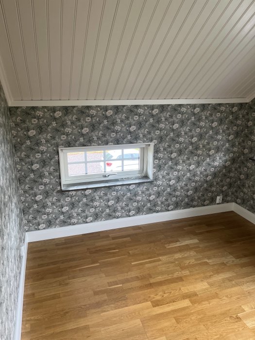 Renoverat rum med trägolv, vägg med blommigt tapet och snedtak med vita paneler.