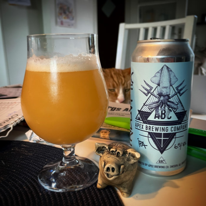 Ölglas med grumlig, gyllene dryck framför ölburk från Apex Brewing, nyckelpigafigur och en katt i bakgrunden.