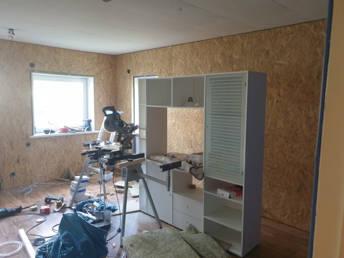 Rum under renovering med OSB-skivor på väggar, verktyg på golv och en vit hylla.