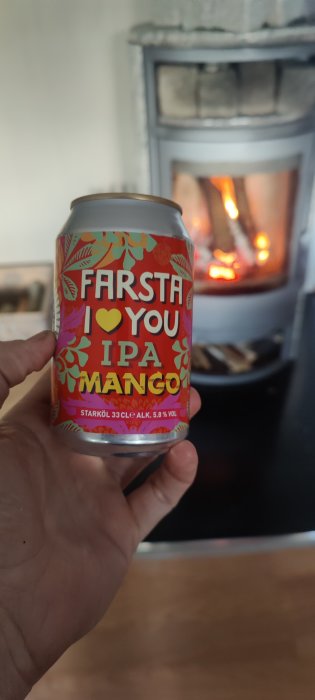 Hand håller en ölburk med texten "FARSTA I LOVE YOU IPA MANGO" framför en brinnande eldstad.