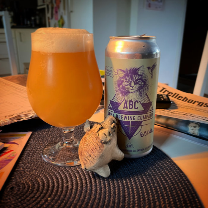 Ett ölglas fyllt med skummande öl bredvid en ölburk från Apex Brewing och en liten figur av en katt.