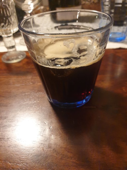 Ett glas fyllt med mörk julmust på ett träbord, halvt drucket med skum kvar på ytan.