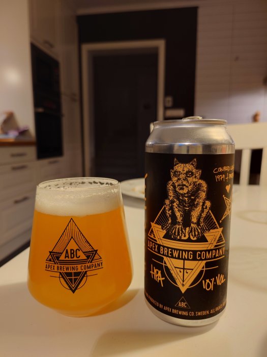 Ölburk och glas märkt "Apex Brewing Company" med öl inuti, står på ett köksbord.