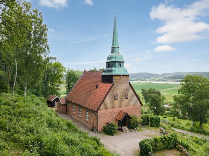 Gammal kyrkobyggnad omgiven av grönska, omvandlad till bostad, med synliga tegeltak och ett högt kyrktorn.