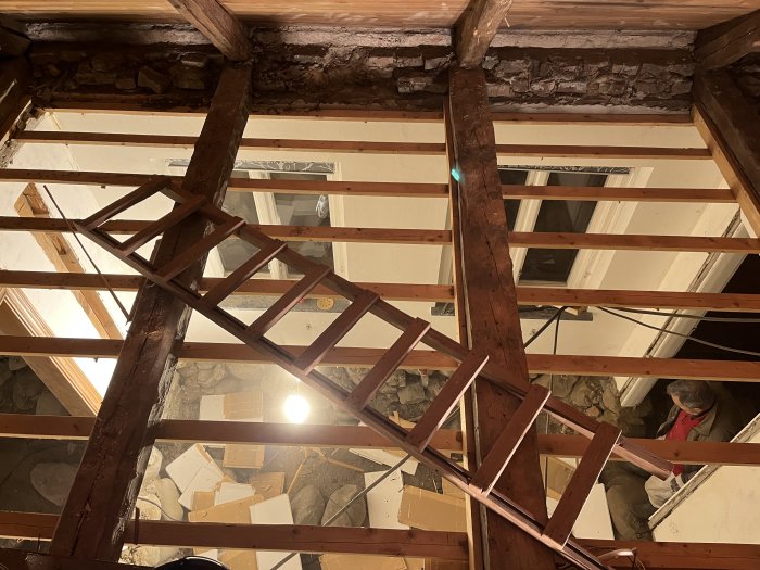 Vy uppåt mot ett hus takstomme under renovering, träbjälkar och en stege synlig, byggmaterial på golvet.