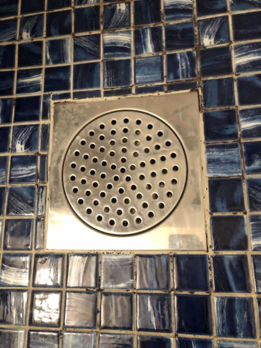Rostfritt golvavlopp med perforerad lockplatta på blå mosaikfliser i en dusch.