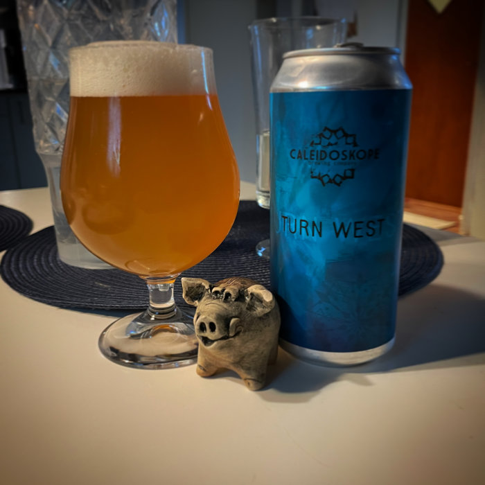 Ett glas med öl och en humleburk märkt "TURN WEST" bredvid en liten grisfigur på ett bord.