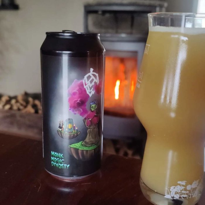 Ölburk med design av munk och magisk rök vid brasa, bredvid ett fullt glas med grumlig öl.