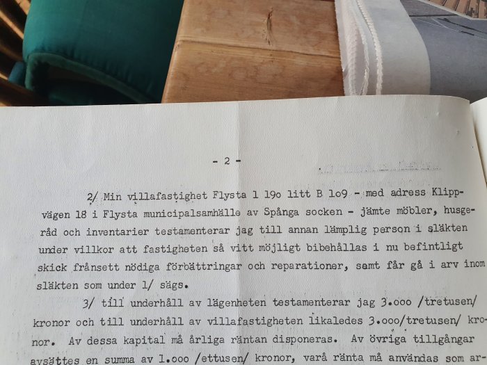 Öppen bok med text om testamente av villa och innehåll på ett träbord framför en grön kudde.