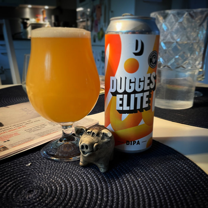 Ett glas öl bredvid en burk märkt "Dugges Elite" och en liten grisskulptur på ett blått underlägg.