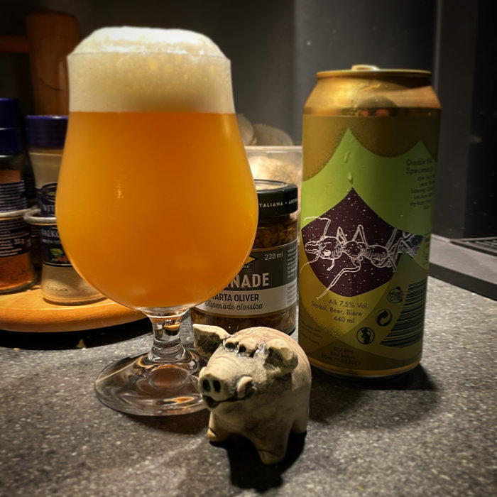Ölglas med skummande öl, burk och en liten griskeramikfigur på ett köksbänk.