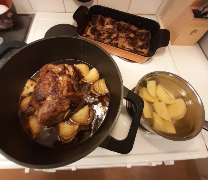 Färdigstekt fläskkarre med frukt i stekgryta, bakform med äpplepaj och kastrull med kokt potatis på spis.