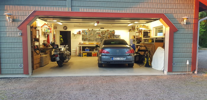 Upplyst garage med öppen skjutport som visar bil, motorcykel och verktyg på väggarna, och golvvärme system.