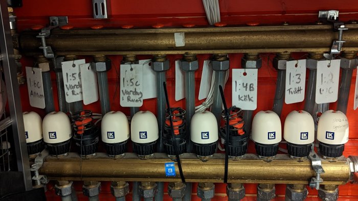 Radiatorventiler på en fördelningsrör med vita markeringsetiketter och synlig sprickbildning.