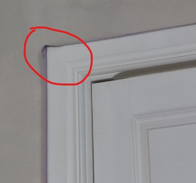 Översta hörnet av en vit dörr och dess karm där skuggor skapar en illusion av försänkning i väggen.