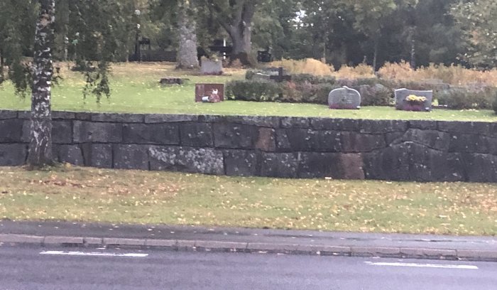 Gråsliten stenmur intill en gräsmatta och gata med träd och gravstenar i bakgrunden.