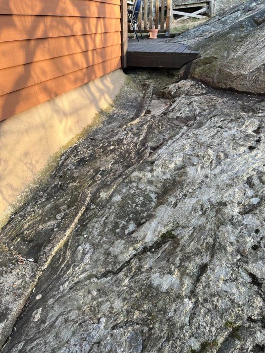 Bild av en naturlig stenformation vid husgrund bredvid trätrall och staket.