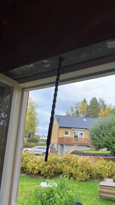 Lång betongborr hängande framför ett fönster med utsikt över en trädgård och hus.