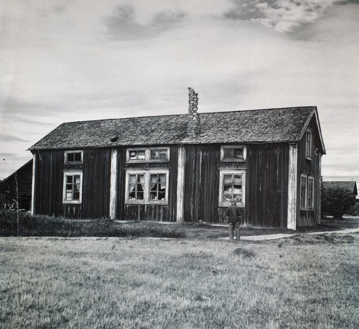 Svartvit bild på ett gammalt trähus med en person som står framför det.