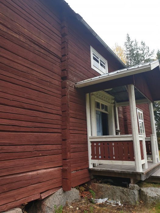 Rödfärgat trähus med hörnklossar och veranda, visar husets exteriör och trädetaljer.