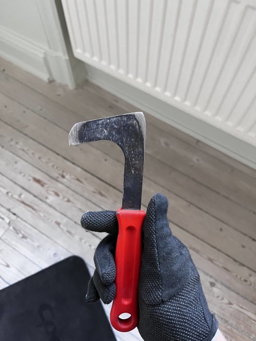 En hand i en svart arbetsvante håller en sliten spackelspade med rött handtag.