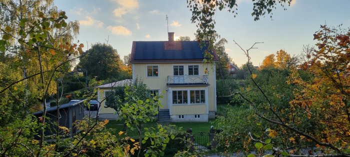 Ett gult hus med vit veranda och solceller på taket omgivet av grönskande träd i ett bostadsområde i skymning.