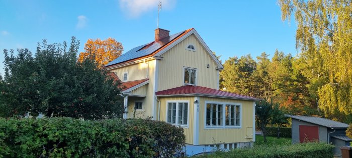 Gult hus med solcellspaneler på taket, omgivet av grönska och träd i solljus.