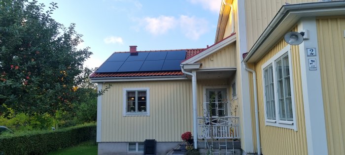 Gult hus med solcellspaneler på taket, trädgård och fruktträd i förgrunden.