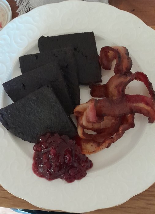 Vit tallrik med blodpudding, knaperstekt bacon och lingonsylt på ett träbord.