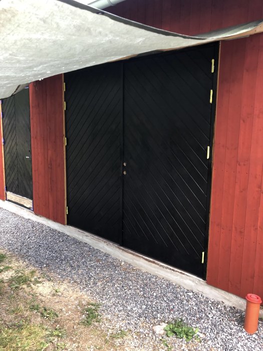 Nyinstallerade, svarta garagedörrar på rödmålad byggnad under presenningstak.