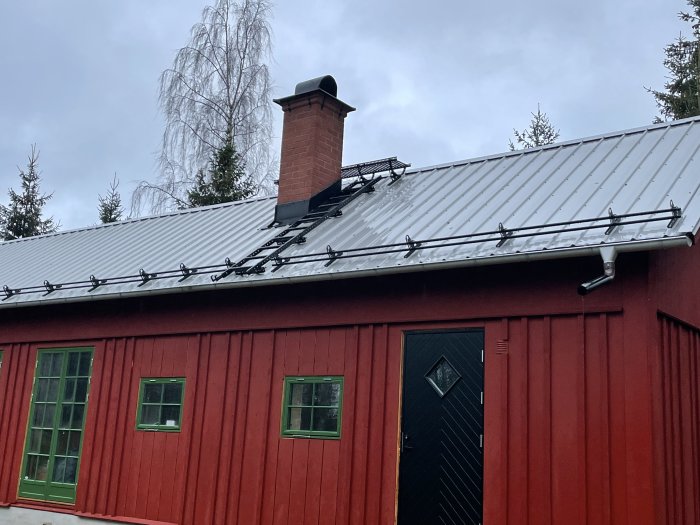 Rött trähus med silverfärgat plåttak utrustat med svarta snörasskydd och skorstensmurskydd, samt takstege.