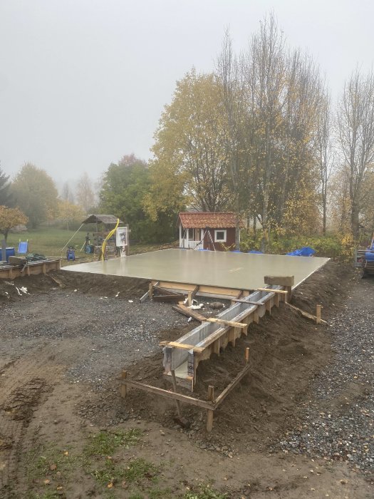 Nyligen gjuten betongplatta med formar och tom yta redo för golvränna, omgiven av byggmaterial och grått väder.