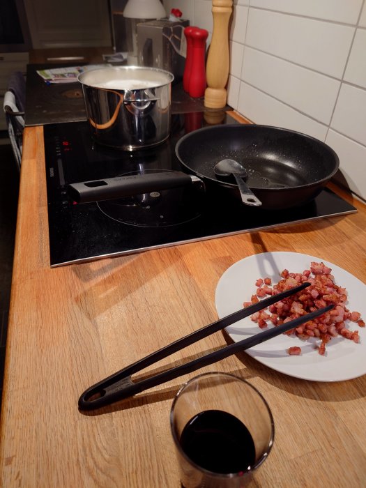 Köksbänk med kokande kastrull, stekpanna, tallrik med baconbitar och glas med rödvin.