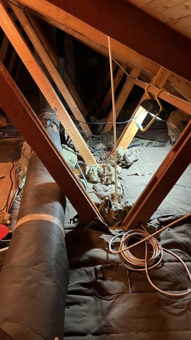 Rörigt vindsutrymme under renovering med isoleringsmaterial, träreglar och ett stort ventilationsrör.