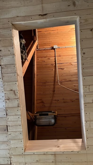 Vindslucka i taket med öppet hål, råspontklädda sidor, och synlig arbetsbelysning i en träkonstruktion.