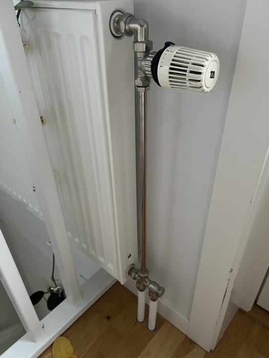 Väggmonterat radiatorlement med termostatventil och synliga rörledningar mot en vit vägg.