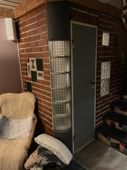 Hörn med glassblocksvägg och stängd dörr i rödteglad källare, möblerad med fåtölj och kuddar.