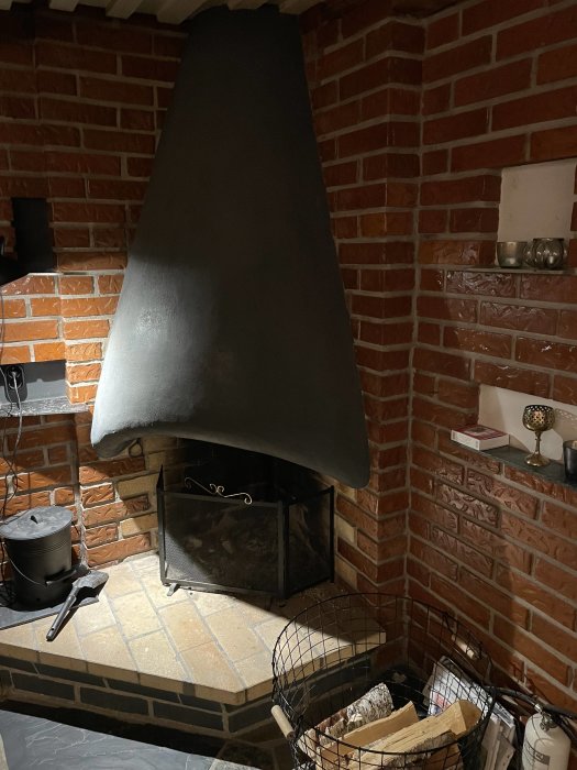 Öppen spis med röd tegelvägg och svart spiskåpa i en källare, inklusive vedkorg och eldstadsredskap.
